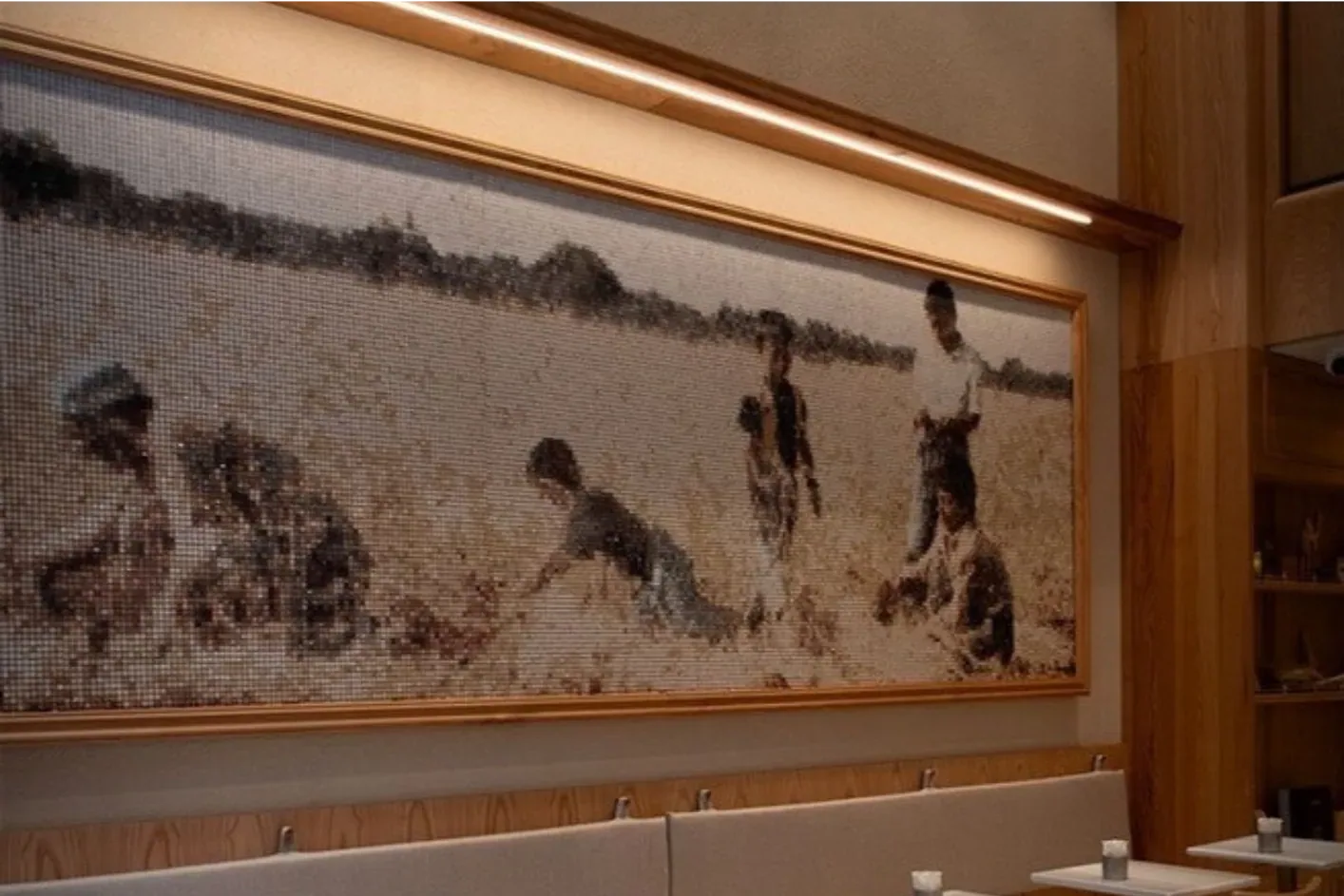 A Mosamia mosaic running the length of a café wall above a banquette, the panoramic scene worked in warm stone tesserae inside an oak frame and lit by a slim cove light above.