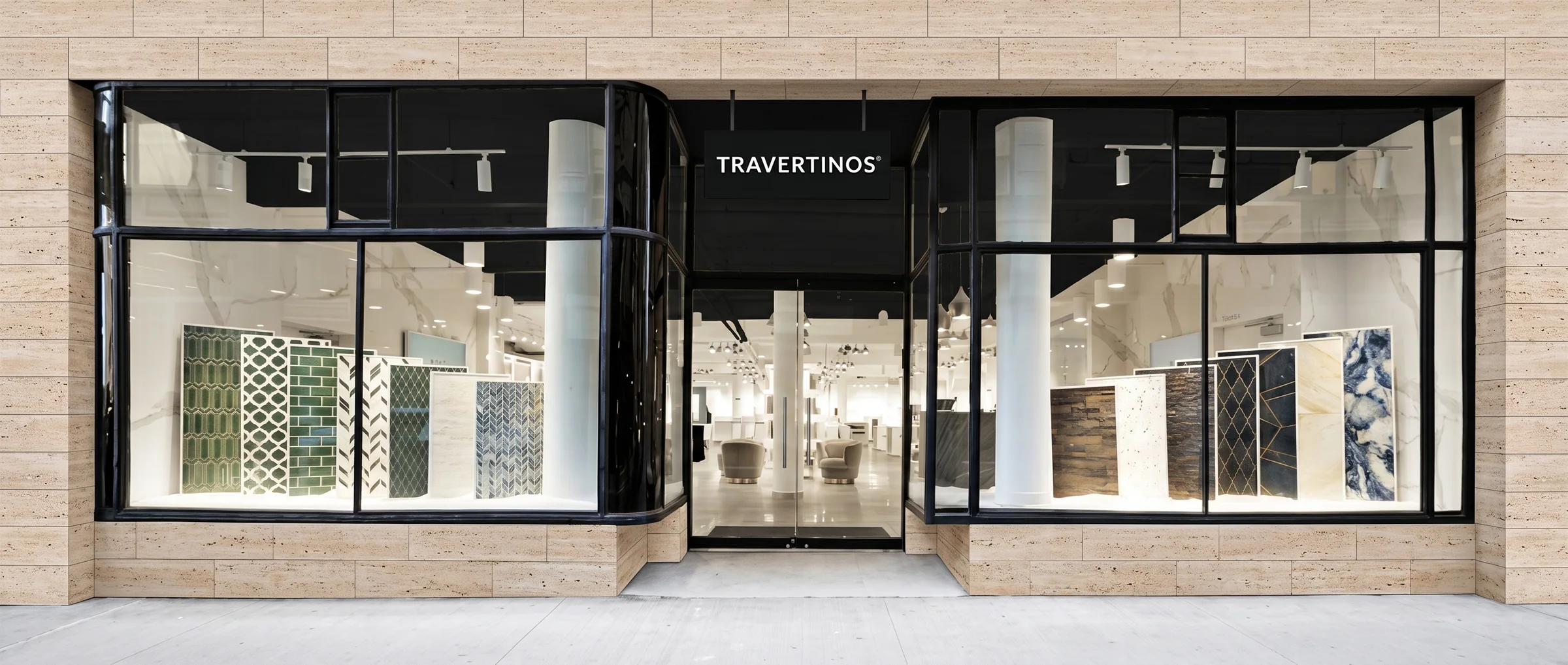 The Travertinos showroom shot square-on: a travertine-clad frontage with black steel-framed glazing either side of a recessed entrance, tile and slab display panels ranged inside under track lighting.