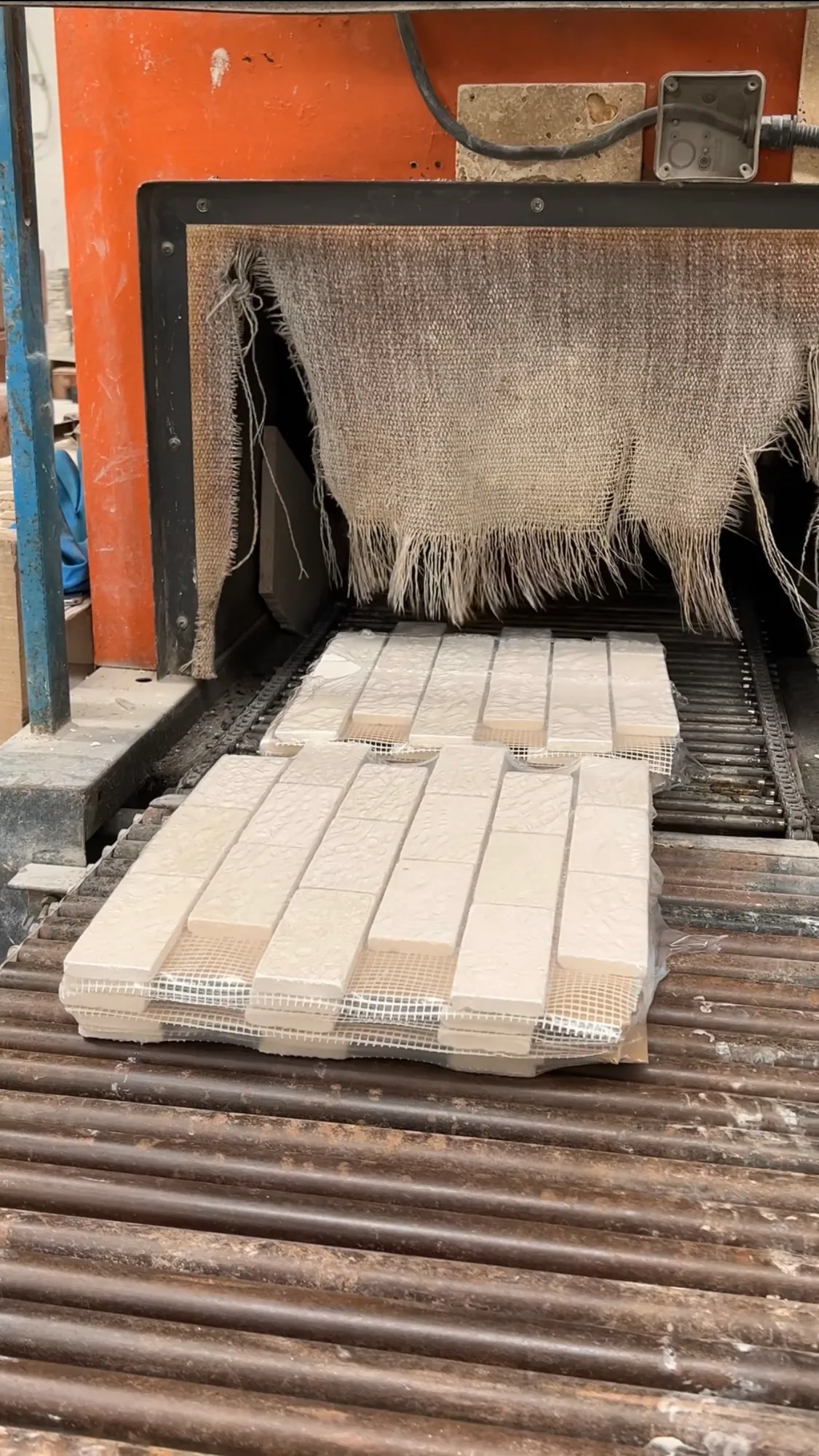 Mesh-backed stone panels on a roller conveyor entering a shrink-wrap tunnel, the machine's frayed heat curtain hanging across its mouth.