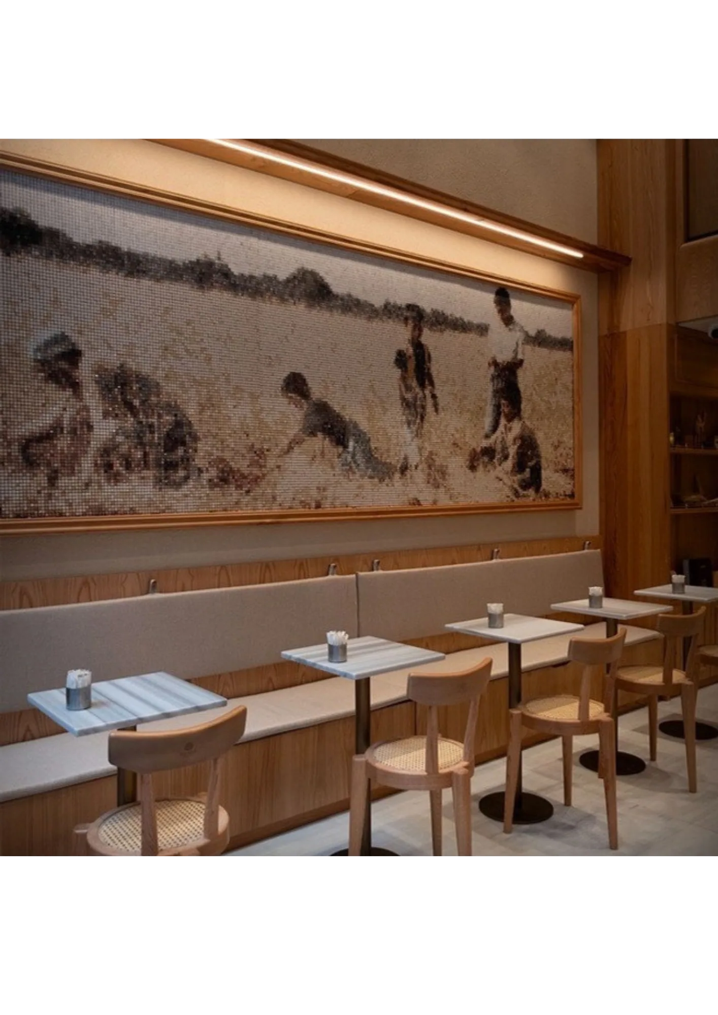 The finished mosaic photographed in place: the panoramic panel in a light timber frame running the length of a café wall above banquette seating, lit by a warm linear fixture, with marble-topped tables and cane chairs below.